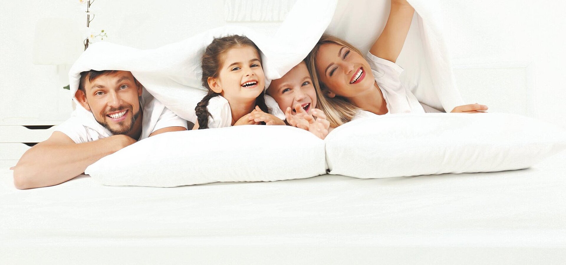 Eine Familie aus Vater, Mutter und zwei Kindern liegt komfortabel und fröhlich auf weißen Kissen unter einer weißen Bettdecke. Das Bild strahlt Gemütlichkeit und Gelassenheit aus.