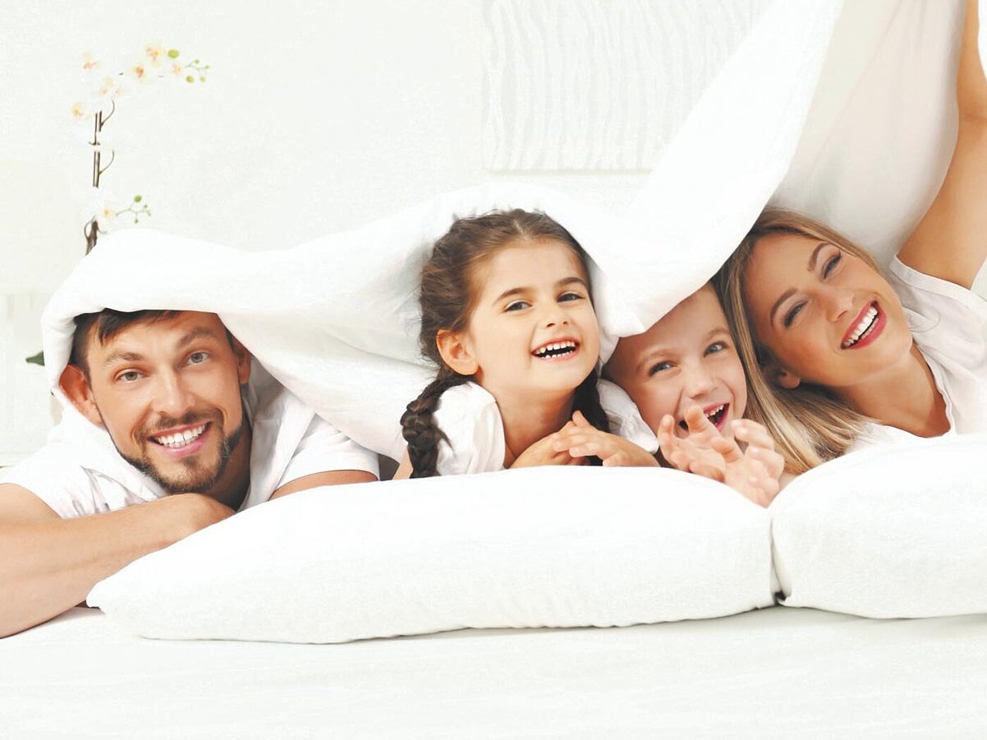 Eine Familie aus Vater, Mutter und zwei Kindern liegt komfortabel und fröhlich auf weißen Kissen unter einer weißen Bettdecke. Das Bild strahlt Gemütlichkeit und Gelassenheit aus.
